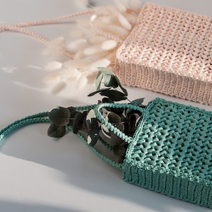 May include: Two handmade woven square handbags. One is light pink, the other is teal. Both have woven straps. The teal bag is open and contains green leaves. The bags are on a white surface with dried flowers.