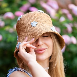 Puede incluir: Una mujer con un sombrero de paja con adornos de flores azules. El sombrero le cubre los ojos y ella sonríe. Lleva un vestido de rayas azules y blancas.