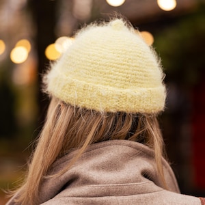 Può includere: Un cappello di maglia giallo chiaro e soffice è indossato da una persona, con lunghi capelli biondi che scendono. La persona indossa un cappotto beige. Lo sfondo è sfocato con luci bokeh.