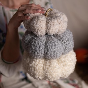 May include: A stack of three crocheted pumpkins in white, gray, and cream. The top pumpkin has a gold loop handle.