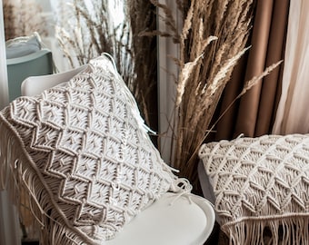 May include: Two cream-colored macrame pillows with fringe trim. The pillows are on a white chair and a brown surface.