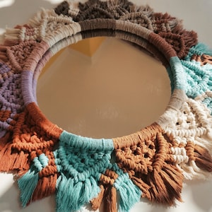 May include: A round mirror with a macrame frame in shades of brown, beige, white, teal, and purple. The frame has a fringe detail around the edge.