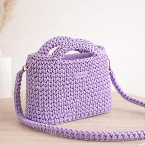 Può includere: Una borsa all'uncinetto color lavanda con manico e tracolla staccabile. La borsa ha una forma rettangolare e una superficie strutturata. La borsa è appoggiata su un tavolo di legno chiaro.