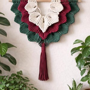 Peut inclure: Suspension murale en macramé à plusieurs niveaux, dans les tons de vert, bordeaux et crème, suspendue à une cheville en bois. Le design présente un bord festonné et une longue gland bordeaux. Le macramé est exposé sur un mur blanc, entouré de plantes en pot.