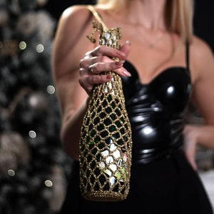 以下が含まれることがあります： メッシュのデザインが施されたゴールドのボトルカバー。ボトルはメッシュ越しに一部見えています。カバーは、黒い革のトップを着た人が持っています。