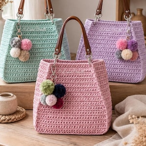 May include: Three crochet tote bags in mint green, lavender, and soft pink. Each bag features a structured trapezoidal shape, brown leather-like handles attached with silver metal rings, and a decorative cluster of four small yarn pom-poms.
