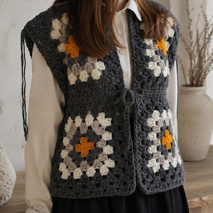 May include: A sleeveless crochet vest constructed from dark gray, light gray, and white granny squares with vibrant orange centers. The vest features a front tie closure, a V-neckline, and visible yarn fringe details along the shoulder seams.
