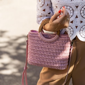 以下が含まれることがあります： 木製のハンドルと取り外し可能なピンクのショルダーストラップが付いた、ダスティローズのクロシェ編みハンドバッグ。白いクロシェ編みトップスとベージュのパンツを履いた人がバッグを持っています。バッグの質感がわかります。