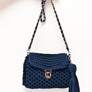 Puede incluir: Un bolso de hombro de punto azul oscuro con una correa de cadena plateada y un borla. El bolso tiene un cierre de solapa con un broche plateado.