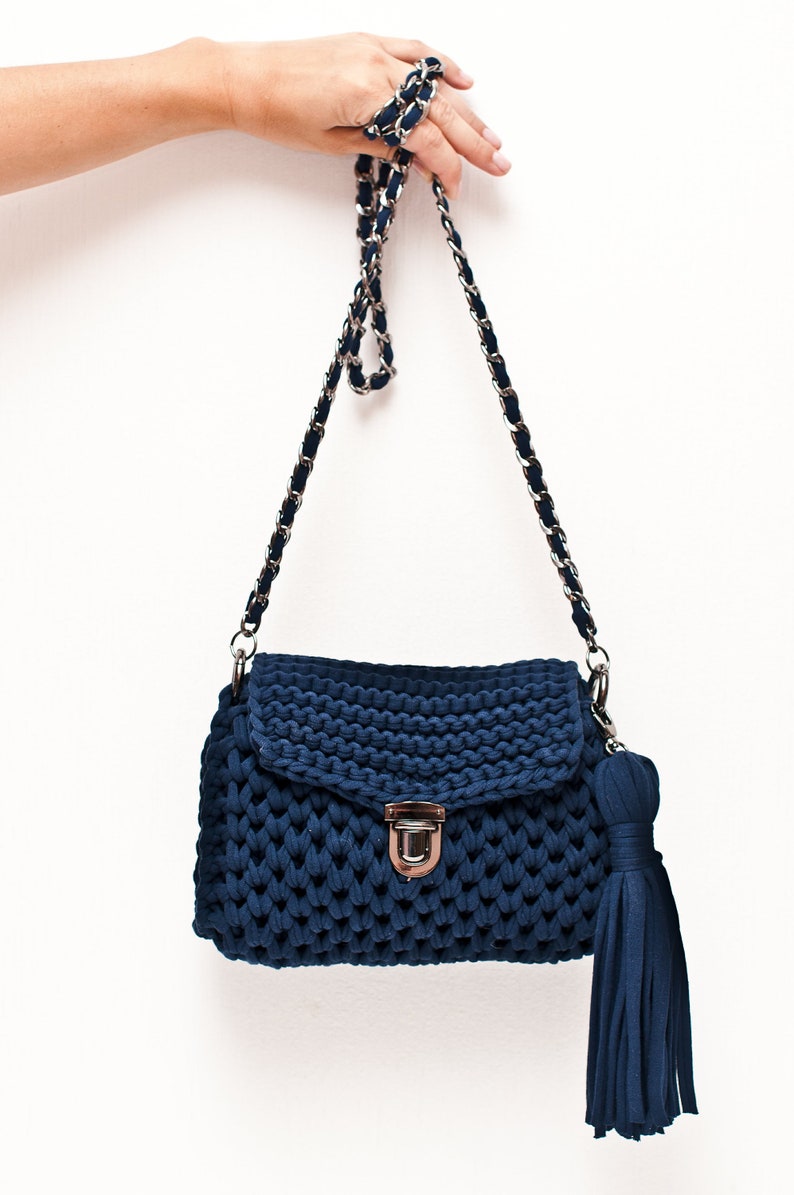 Puede incluir: Un bolso de hombro de punto azul oscuro con una correa de cadena plateada y un borla. El bolso tiene un cierre de solapa con un broche plateado.