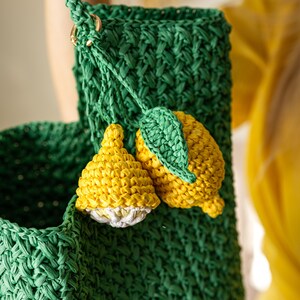 May include: A green crocheted bag with a woven texture, featuring a braided strap and gold-colored hardware. The bag is adorned with two yellow and white crocheted lemon charms with green leaves, adding a playful touch to the design.