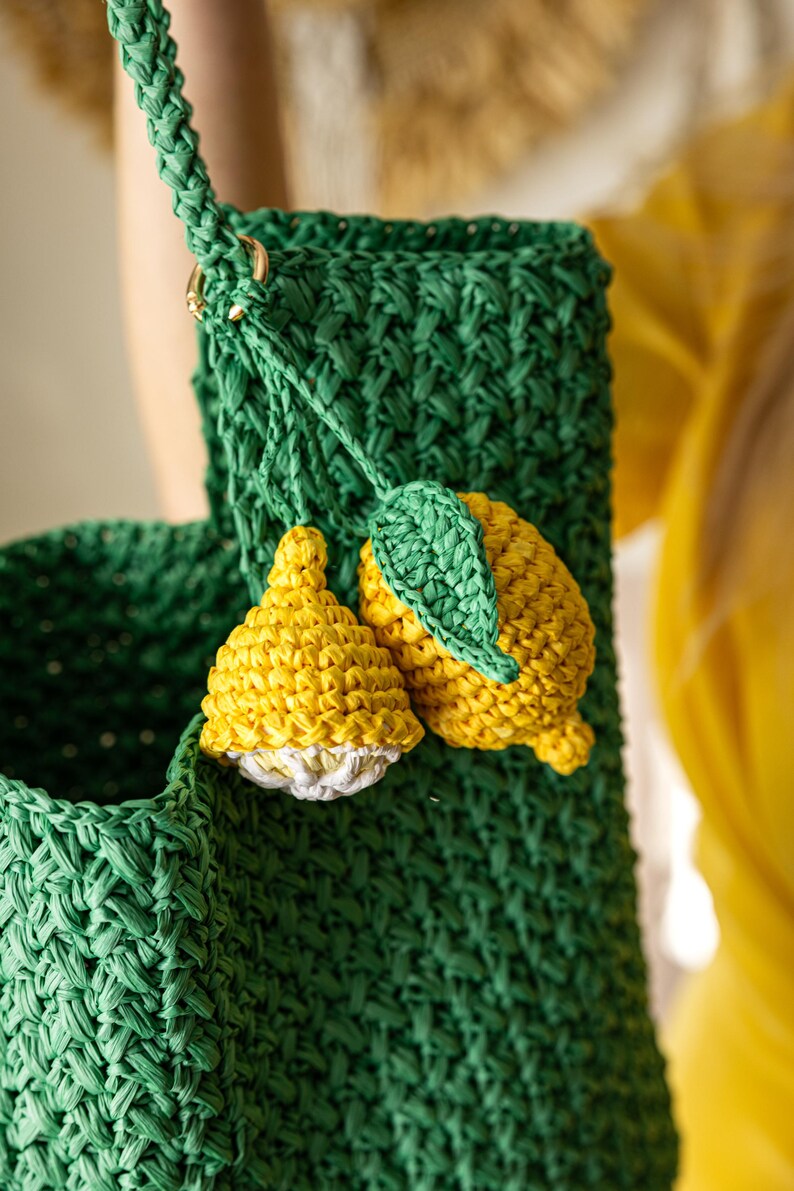 May include: A green crocheted bag with a woven texture, featuring a braided strap and gold-colored hardware. The bag is adorned with two yellow and white crocheted lemon charms with green leaves, adding a playful touch to the design.
