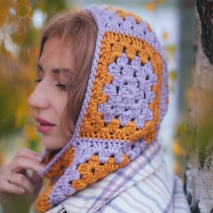 May include: A handmade crochet hood in a granny square pattern. The hood features a square design in shades of lavender and mustard yellow. The hood is worn over a plaid shirt.