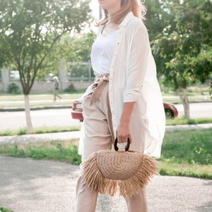 May include: A woman wearing sunglasses, a white shirt, and beige pants, carrying a woven straw handbag with a fringe. The bag has a wooden handle. The outfit is light-colored, suitable for a sunny day.