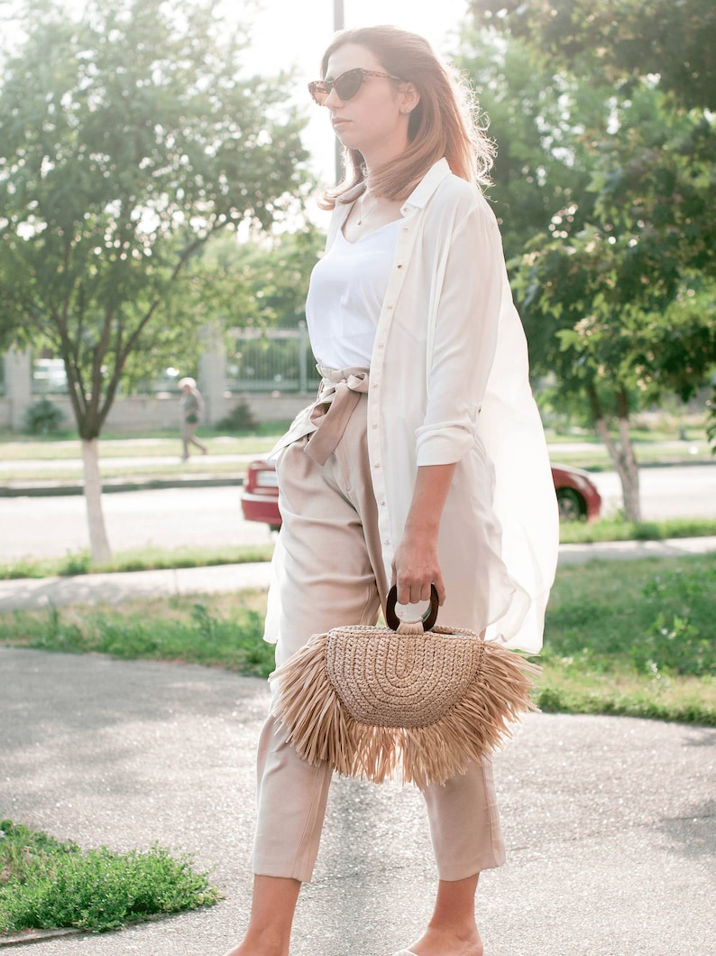 May include: A woman wearing sunglasses, a white shirt, and beige pants, carrying a woven straw handbag with a fringe. The bag has a wooden handle. The outfit is light-colored, suitable for a sunny day.