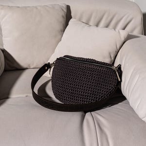 May include: A dark gray crocheted fanny pack with a black leather strap. The bag is sitting on a white couch.
