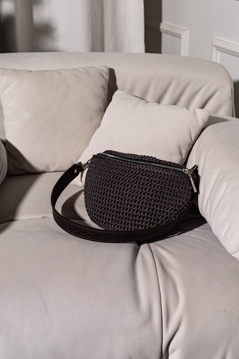 May include: A dark gray crocheted fanny pack with a black leather strap. The bag is sitting on a white couch.