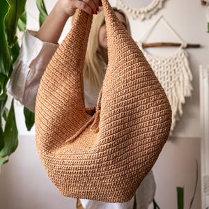 May include: A large, hand-crocheted bag in a warm, tan color. The bag has a distinctive, rounded shape with a drawstring closure. The textured stitch creates a ribbed effect. The bag is held up by a person.