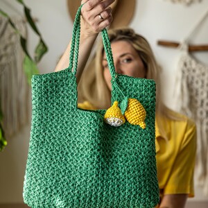 May include: A green crochet tote bag with a rectangular shape and two handles. The bag is decorated with two yellow lemon appliqu&eacute;s. The bag is held up by a person.