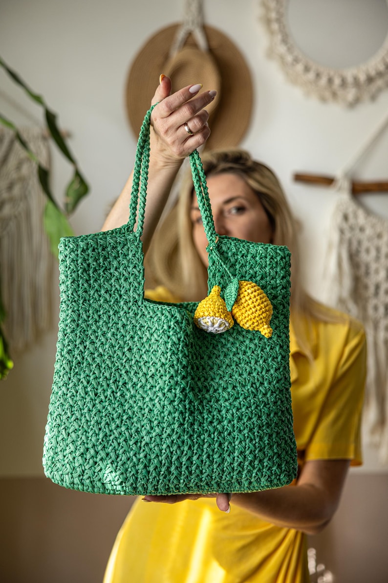 May include: A green crochet tote bag with a rectangular shape and two handles. The bag is decorated with two yellow lemon appliqu&eacute;s. The bag is held up by a person.