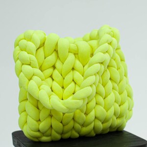 Puede incluir: Un bolso tote de punto amarillo brillante hecho con hilo grueso. El bolso tiene un dise&ntilde;o trenzado y est&aacute; sentado en un taburete de madera negro.