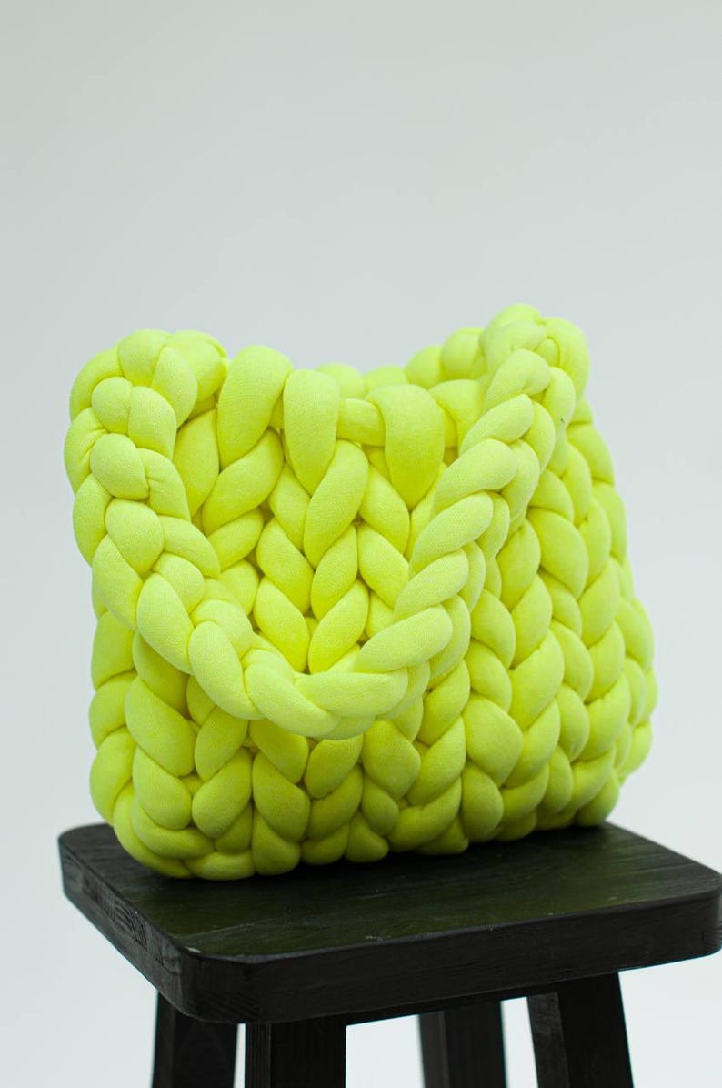 Puede incluir: Un bolso tote de punto amarillo brillante hecho con hilo grueso. El bolso tiene un dise&ntilde;o trenzado y est&aacute; sentado en un taburete de madera negro.