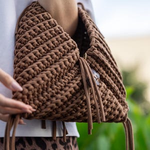 May include: A brown, hand-crocheted bag with a unique, textured design. The bag features a triangular shape and is adorned with long, dangling tassels. A small charm hangs from the side. The bag is held against a blurred background.
