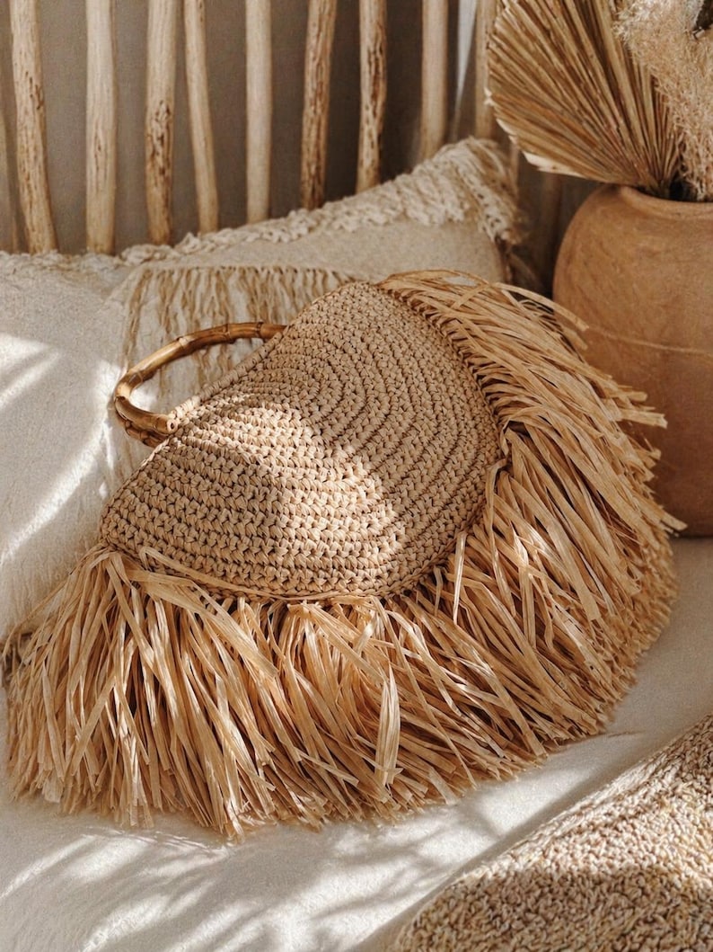 May include: A beige woven handbag with a circular design and a bamboo handle. The bag features long, fringed edges, adding a textured, bohemian style. The bag is set against a soft, neutral background, suggesting a natural, earthy aesthetic.