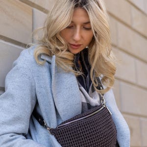 May include: A woman wearing a light blue coat with a brown crocheted fanny pack worn across her waist.