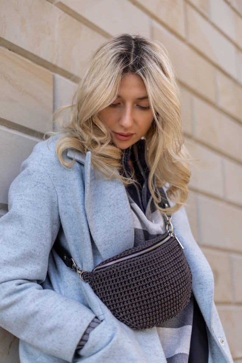 May include: A woman wearing a light blue coat with a brown crocheted fanny pack worn across her waist.