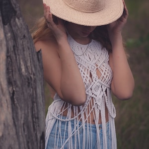 Peut inclure: Une femme portant un haut en crochet blanc avec des d&eacute;tails en frange et un jean bleu. Elle tient un chapeau de paille &agrave; larges bords devant son visage.