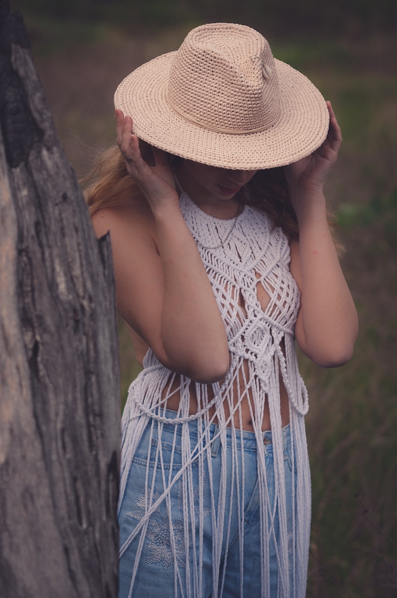 Peut inclure: Une femme portant un haut en crochet blanc avec des d&eacute;tails en frange et un jean bleu. Elle tient un chapeau de paille &agrave; larges bords devant son visage.