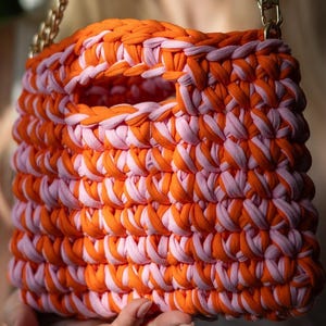 May include: A handmade, square handbag in orange and light pink. The bag is made from thick, woven fabric strips, with a handle opening and a gold chain strap. The bag is held by a person. The bag's dimensions are not visible.