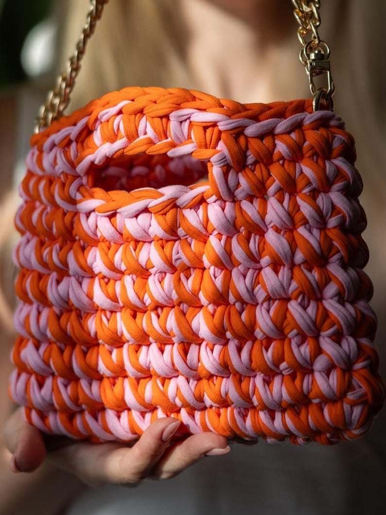 May include: A handmade, square handbag in orange and light pink. The bag is made from thick, woven fabric strips, with a handle opening and a gold chain strap. The bag is held by a person. The bag's dimensions are not visible.