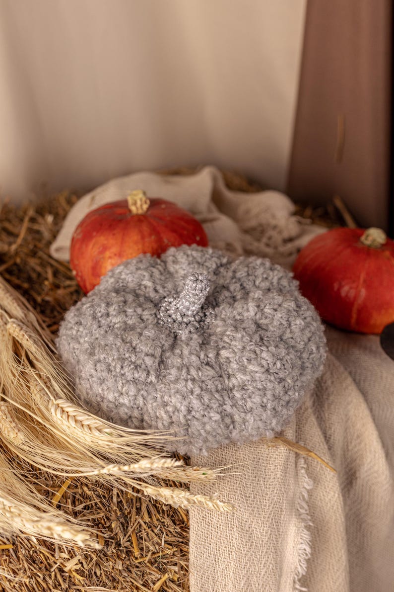 May include: A gray fuzzy pumpkin-shaped hat sits on a bed of straw with two red pumpkins beside it.