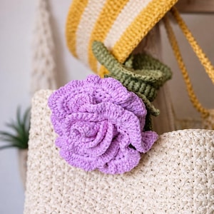 May include: A handcrafted, light purple crochet flower with green leaves rests on a woven bag. A yellow and cream striped crochet hat with a long strap is visible in the background. The items are part of a collection of handmade accessories.