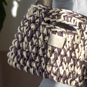 May include: A cream and grey woven handbag with a geometric design. The bag has a strong handle and a silver-tone chain strap. A small rectangular tag with text is attached to the front.