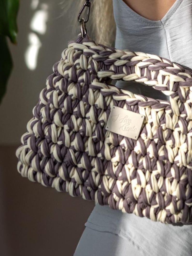 May include: A cream and grey woven handbag with a geometric design. The bag has a strong handle and a silver-tone chain strap. A small rectangular tag with text is attached to the front.