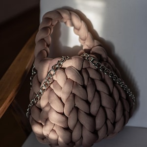 Puede incluir: Un bolso de mano de punto marr&oacute;n claro con una correa de cadena plateada. El bolso est&aacute; hecho de tela gruesa y tejida y tiene una forma redonda ligeramente aplanada.
