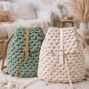 Op de afbeelding: Twee handgemaakte gehaakte rugzakken. Eén is saliegroen, de andere is crèmekleurig. Beide hebben een schubachtige textuur, leren riemen en een gespsluiting. De tassen liggen op een neutraal getint tapijt.