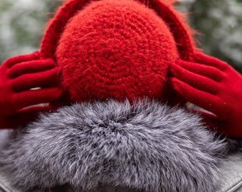May include: A close-up of a winter outfit featuring a bright red knitted earmuffs and matching gloves. The earmuffs are positioned over a gray fur collar, which is part of a gray jacket. The background is blurred, suggesting an outdoor setting.