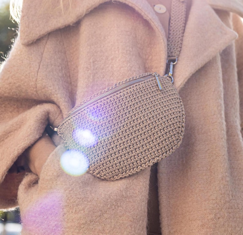 May include: A beige crocheted fanny pack with a silver zipper and a long strap. The bag is worn over a beige coat.