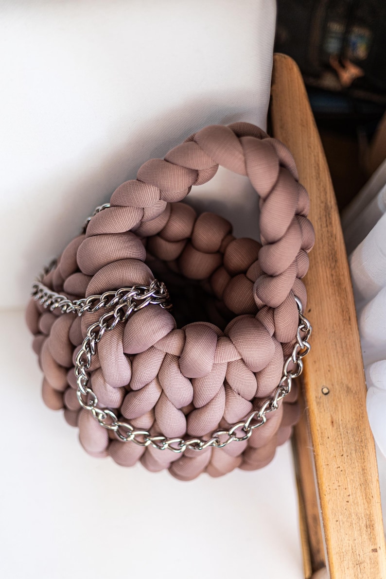 Puede incluir: Un bolso de crochet de color marr&oacute;n claro con una correa de cadena plateada. El bolso est&aacute; hecho de tela suave y tiene un dise&ntilde;o &uacute;nico y texturizado.