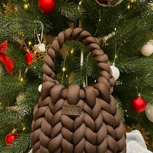 Puede incluir: Un bolso de mano de punto marr&oacute;n con un dise&ntilde;o trenzado y una etiqueta de cuero que dice "LDAR". El bolso est&aacute; delante de un &aacute;rbol de Navidad con adornos rojos y blancos.
