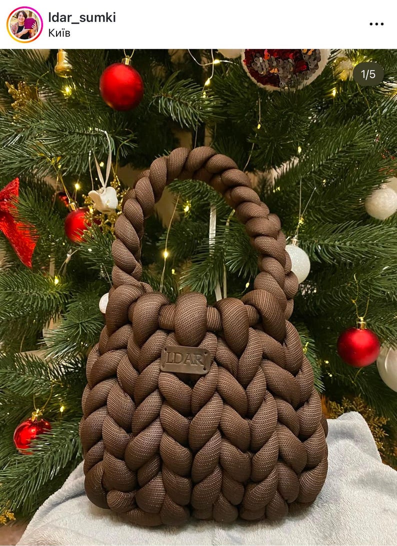Puede incluir: Un bolso de mano de punto marr&oacute;n con un dise&ntilde;o trenzado y una etiqueta de cuero que dice "LDAR". El bolso est&aacute; delante de un &aacute;rbol de Navidad con adornos rojos y blancos.