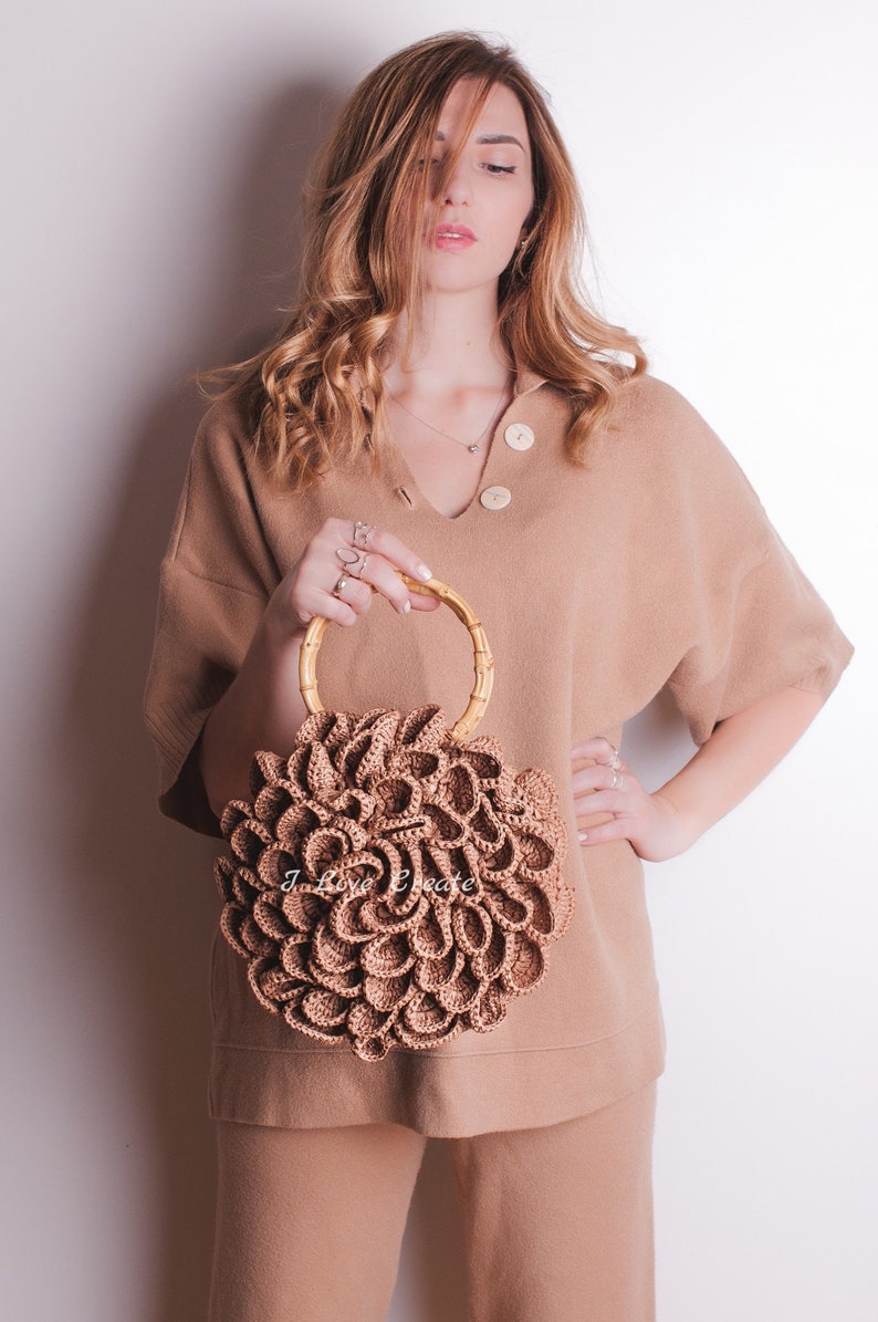 May include: A tan-colored, crocheted handbag with a bamboo handle. The bag has a floral design and is held by a person wearing a tan sweater and pants. The background is a plain white wall.