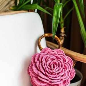 May include: A pink crocheted rose-shaped bag with a bamboo handle. The bag is sitting on a white chair.