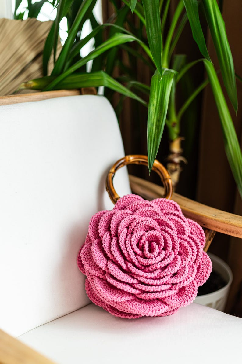 May include: A pink crocheted rose-shaped bag with a bamboo handle. The bag is sitting on a white chair.