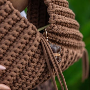 May include: A brown, hand-crocheted handbag with a rounded shape. The bag features a thick, textured stitch and a small, brown leather bear charm with a gold-colored clasp. Long, brown fringe hangs from the side of the bag.