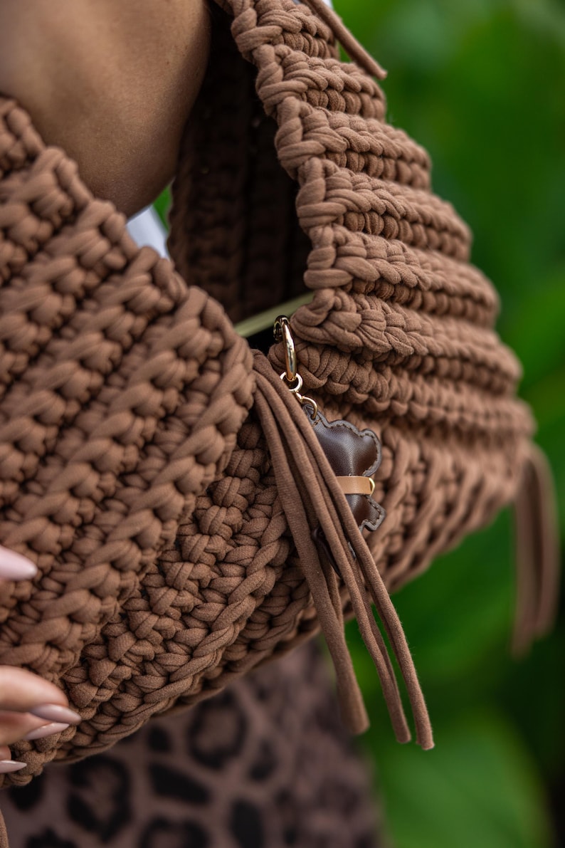 May include: A brown, hand-crocheted handbag with a rounded shape. The bag features a thick, textured stitch and a small, brown leather bear charm with a gold-colored clasp. Long, brown fringe hangs from the side of the bag.